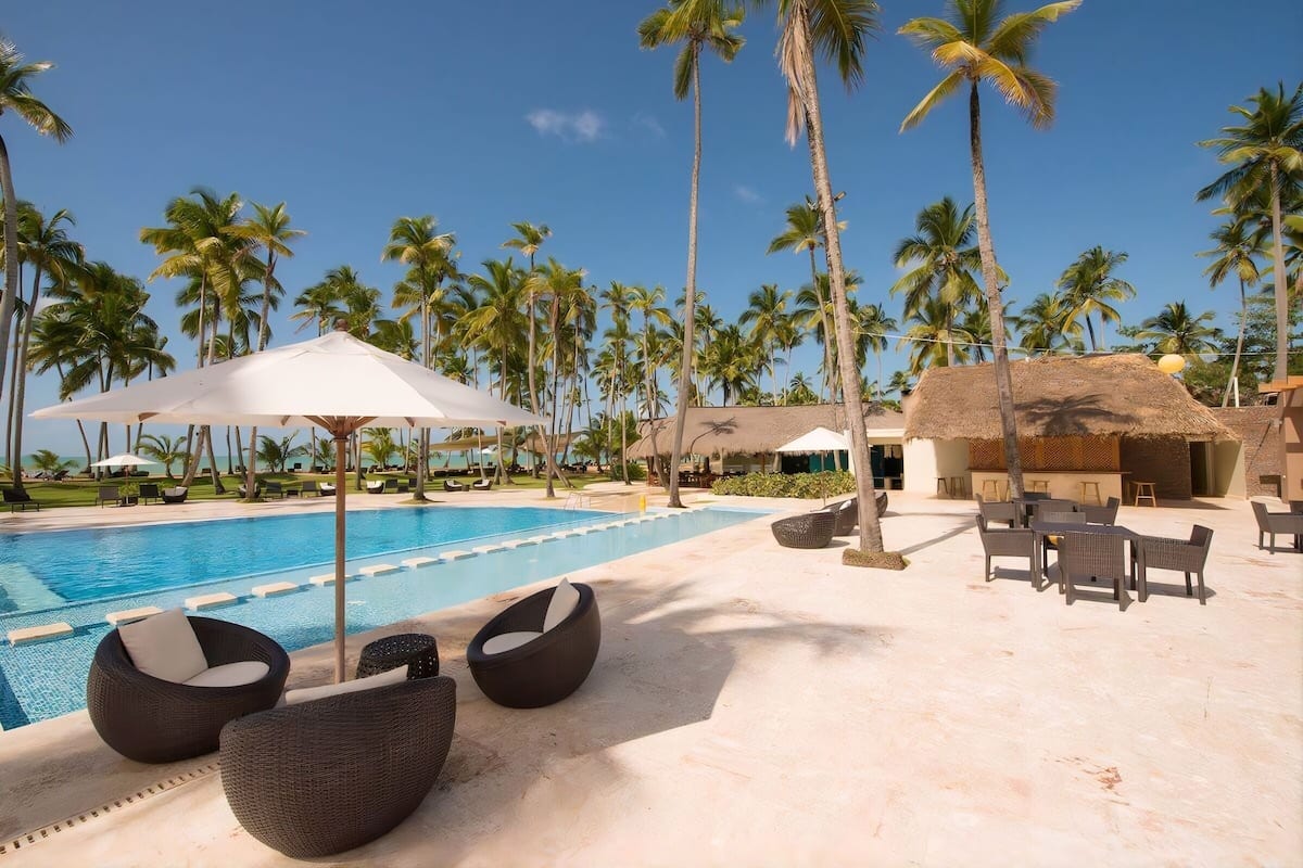 A poolside area with lounge chairs, a thatched-roof structure, and palm trees.