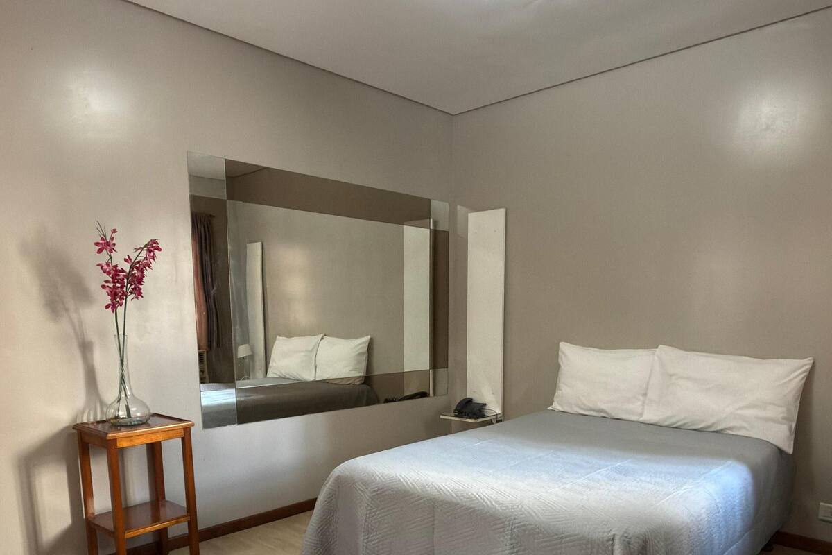 Une chambre à coucher avec un lit, une table de chevet, un miroir et un vase rempli de fleurs.