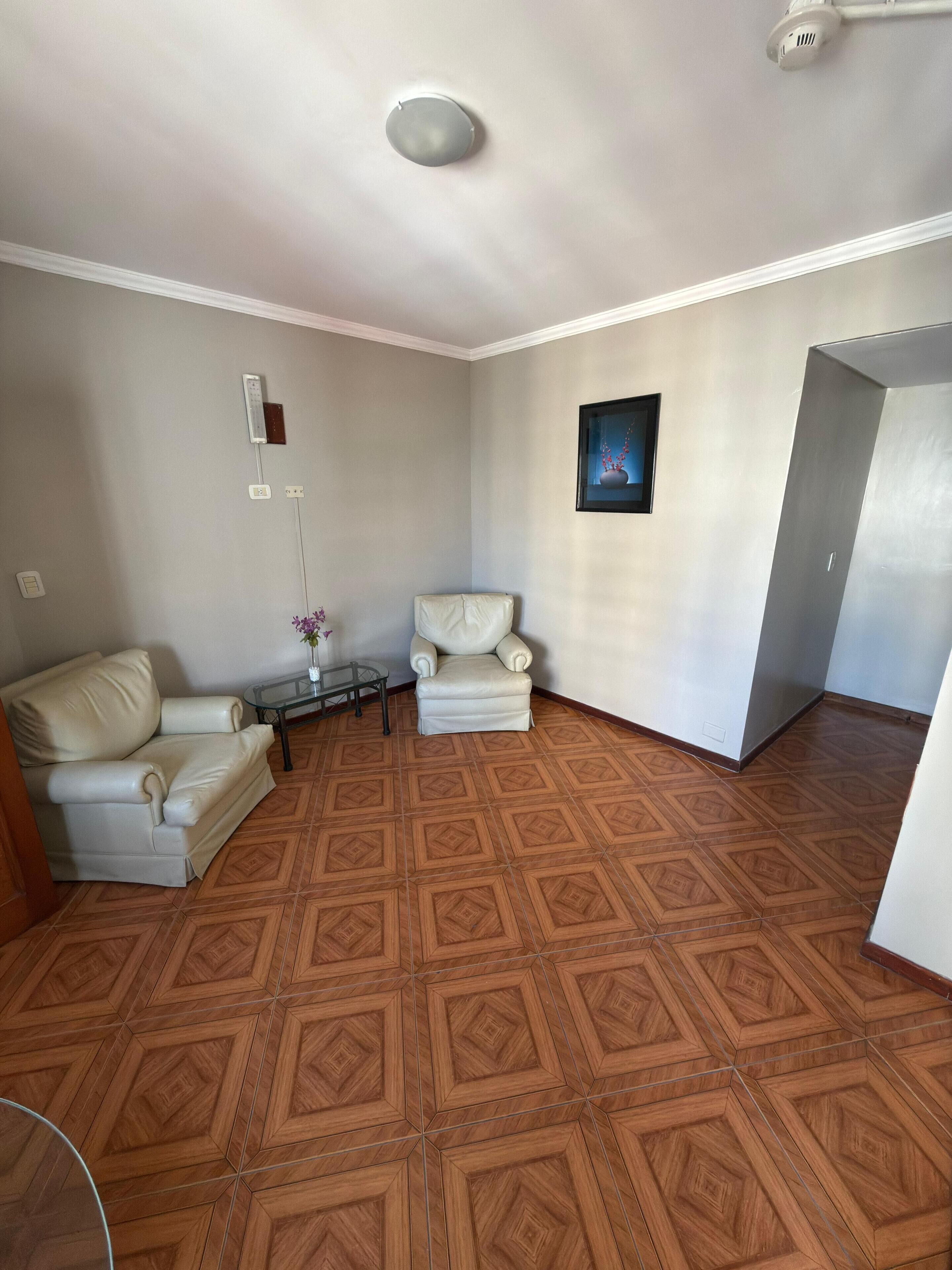 Una habitación con piso estampado, dos sillones blancos, una mesita con un jarrón y un cuadro en la pared.