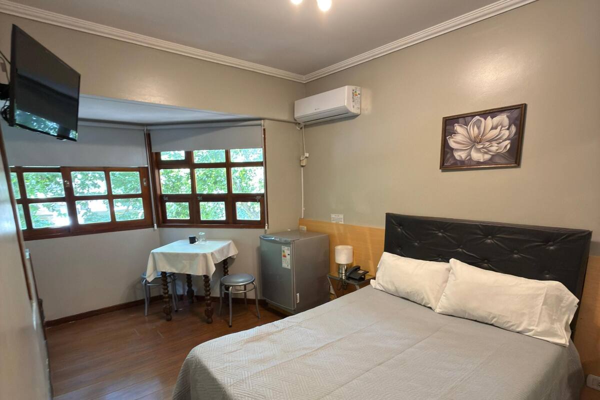 Une chambre d’hôtel équipée d’un lit, d’un bureau, d’une chaise, d’une petite table, d’un climatiseur et d’une fenêtre donnant sur des arbres.