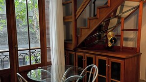 Una habitación con una mesa y sillas de vidrio, una escalera de madera y vistas a través de una ventana con cortinas.
