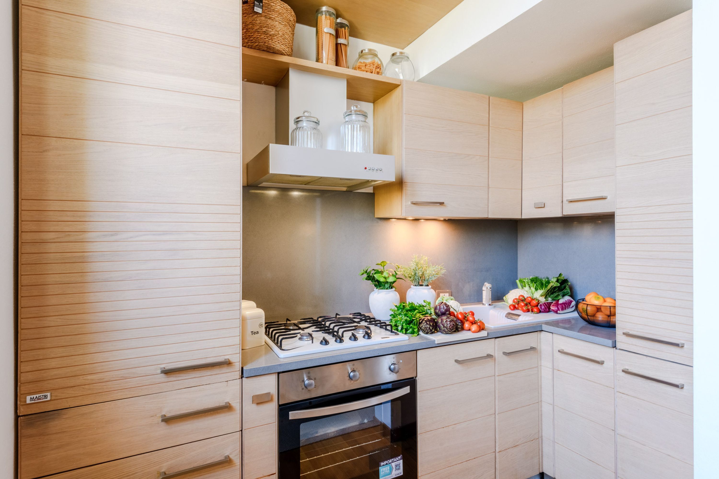 Una cocina moderna con armarios de madera, estufa a gas, horno y una encimera con verduras frescas y frascos.