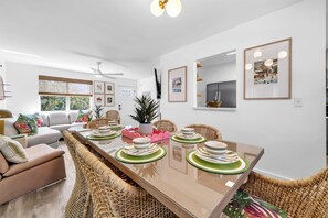 Een moderne eethoek met een lange tafel voor vier personen, omringd door rieten stoelen, een bank en een plafondventilator.