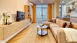 A modern living room with a sofa, coffee table, and TV. There is a dining area with a table and chairs, and a kitchen area with cabinets and a stove. - Soothing 1-bedroom apartment in Floreasca, București (București)
