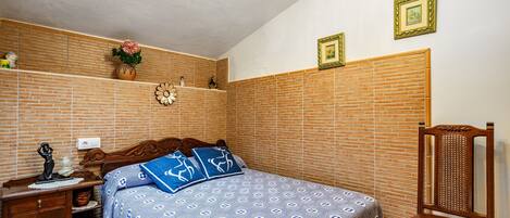 A bedroom with a brick-patterned wall, a wooden nightstand, a bed with patterned bedding, a chair, and a small table with a decorative item.