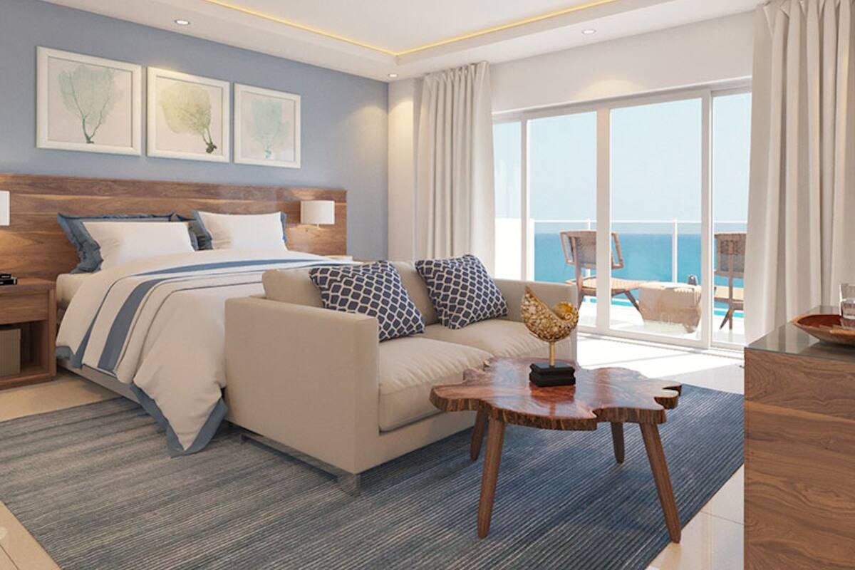 A modern hotel room with a large bed, a sofa, a wooden coffee table, and a view of the sea through large windows.