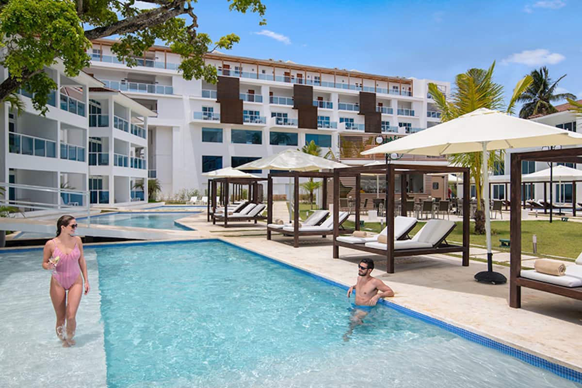 A swimming pool with lounge chairs and umbrellas, a building with multiple balconies, and palm trees.