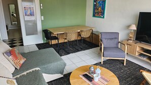Ein Wohnzimmer mit einer Couch, einem runden Couchtisch, einem Esstisch mit Stühlen, einem Fernseher und einem Bild an der Wand.