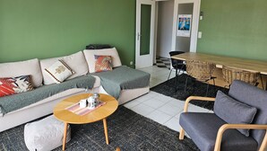 Ein Wohnzimmer mit einem Ecksofa, einem runden Couchtisch, einem grauen Sessel, einem Esstisch mit Stühlen und einer Hängeleuchte aus Weidwerk.