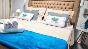 Cama bem arrumada com colcha azul, duas almofadas com a marca 'Villa Biza' e toalhas brancas dobradas sobre a cama.