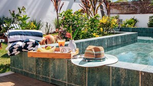 Ambiente à beira da piscina com um deck de madeira, uma toalha, um suporte com comida e bebidas, e um chapéu.