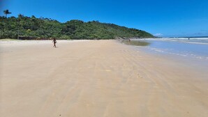 Uma praia com uma pessoa caminhando, céu claro e uma colina arborizada ao fundo.