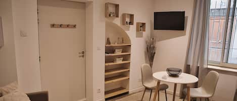 Een kamer met een deur, een klein tafeltje met stoelen, een aan de muur bevestigde televisie en een raam met gordijnen.