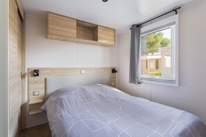 A bedroom with a bed, a wooden headboard, a window with curtains, and a built-in shelf above the bed. - Mobil-Home Mar Estang (Canet-en-Roussillon)