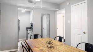 Uma sala de jantar com uma mesa de madeira, quatro cadeiras, uma luminária no teto e uma cozinha ao fundo.