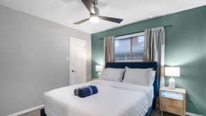 Quarto de hotel com cama, ventilador de teto, mesa de cabeceira com abajur e janela com cortinas.