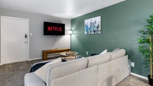 Uma sala de estar com sofá, mesa de centro, TV exibindo 'NETFLIX', um quadro e um vaso com planta.