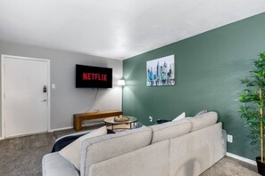 A living room with a sofa, a coffee table, a TV displaying 'NETFLIX,' a painting, and a potted plant. - Arlington 2BR | AT&T, Globe Life, 6 Flags, Parking (Arlington)