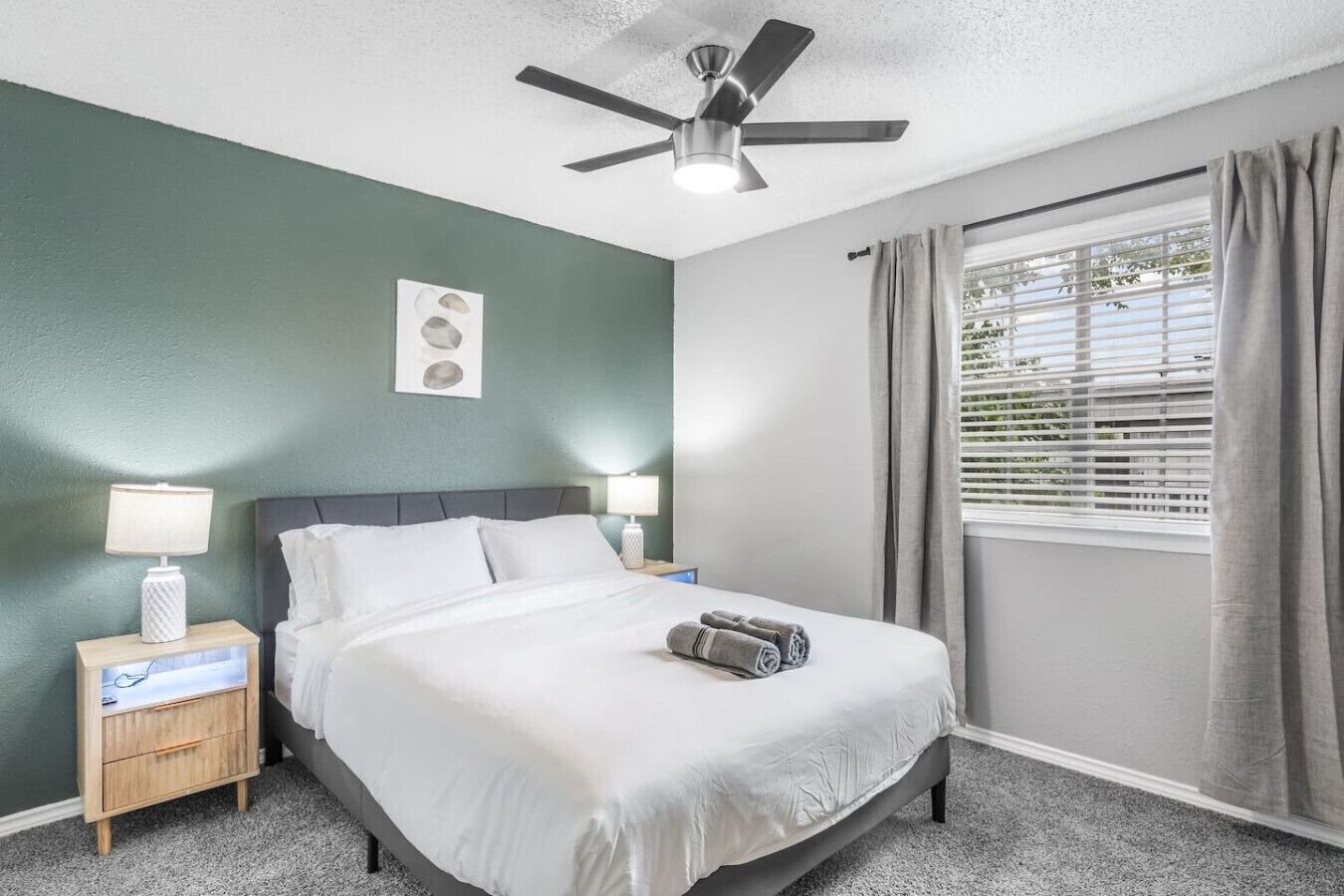 A neatly arranged hotel room with a bed, bedside lamps, a ceiling fan, a window with curtains, and a wall-mounted light fixture.