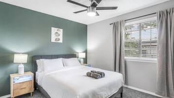 A neatly arranged hotel room with a bed, bedside lamps, a ceiling fan, a window with curtains, and a wall-mounted light fixture.