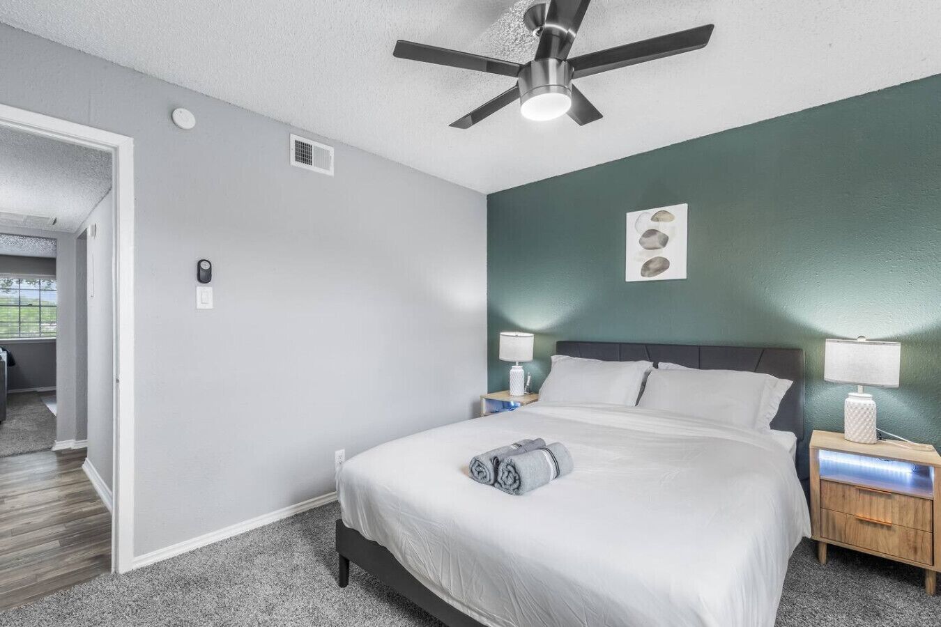 Habitación de hotel con cama, dos mesitas de noche con lámparas, un ventilador de techo, una puerta a otra habitación y un interruptor de luz en la pared.