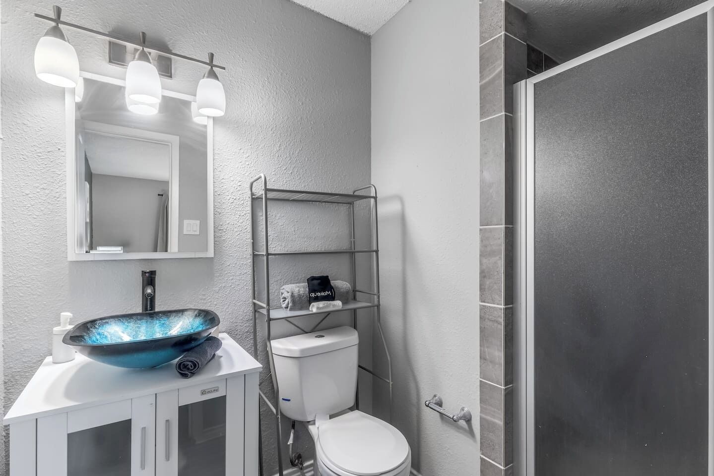 A bathroom with a white sink, a blue vessel sink, a toilet, a metal shelf with towels, and a shower area.