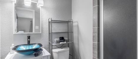 A bathroom with a white sink, a blue vessel sink, a toilet, a metal shelf with towels, and a shower area.