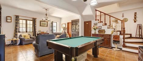 A spacious living room with a pool table, a sofa set, a chandelier, and a staircase leading to an upper level.