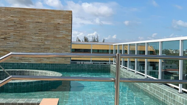 A rooftop pool with clear blue water, a wooden deck, and a modern building with large windows in the background. - Your complete studio apartment in Porto de Galinhas, sun and sea! (Ipojuca)
