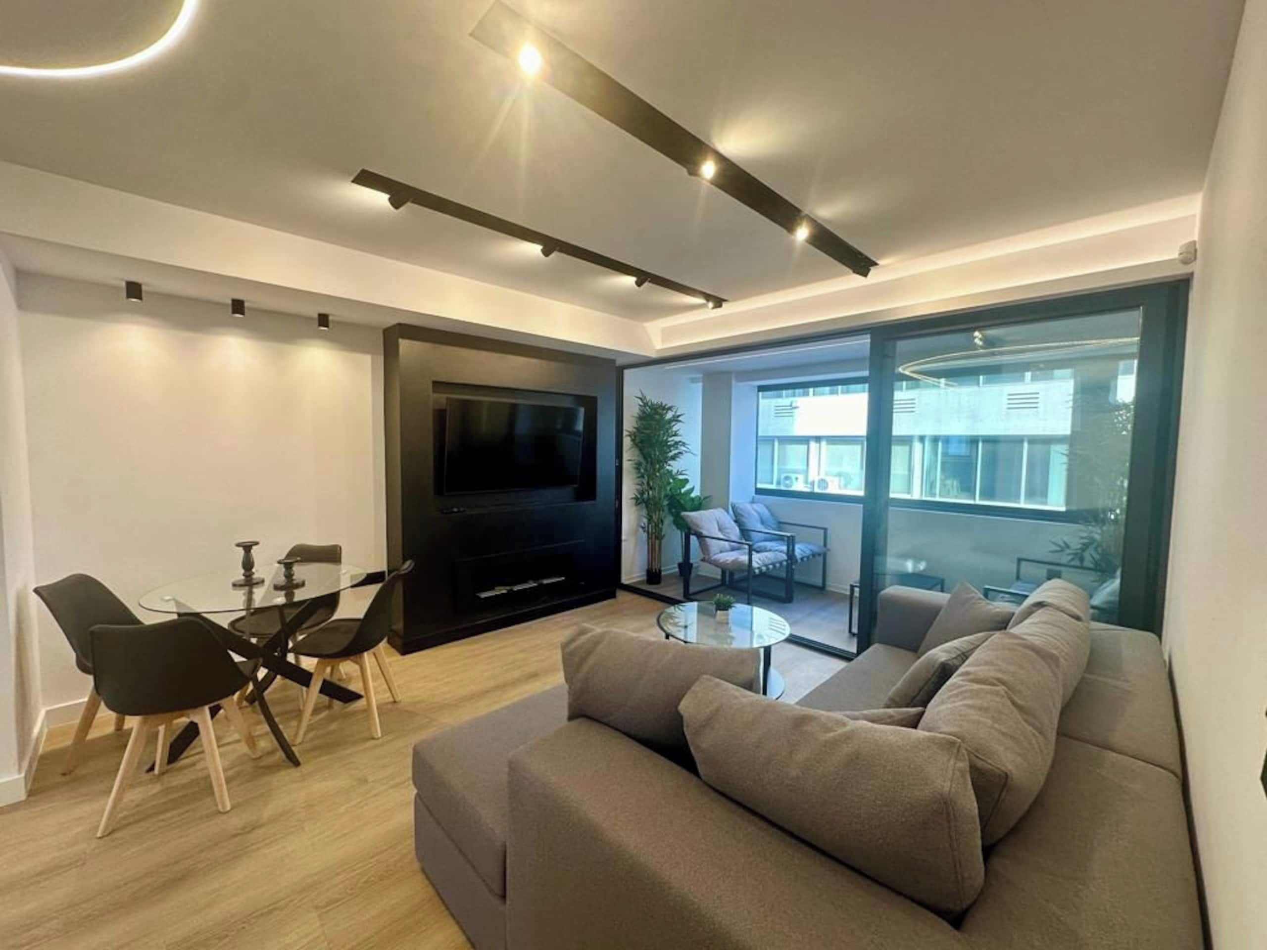 Una sala moderna con televisor de pantalla plana, un sofá, una mesa de comedor con sillas y un ventanal con vistas al exterior.