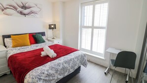 A bedroom with a bed, a nightstand, a lamp, a window with blinds, and a desk with a chair.