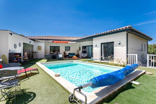 Une piscine recouverte d’une bâche bleue, une terrasse avec des chaises et un bâtiment doté d’un toit en tuiles.