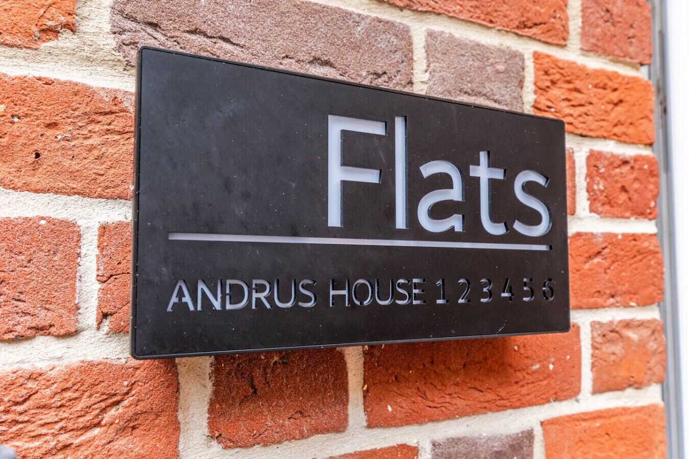 Segno nero con la scritta 'Flats' e 'ANDRUS HOUSE 123456' su un muro di mattoni.