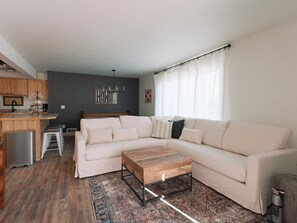 Ein modernes Wohnzimmer mit einer großen, winkelförmigen Couch, einem Holztisch und einer Küchenzeile mit Holzschränken und einem Kühlschrank.