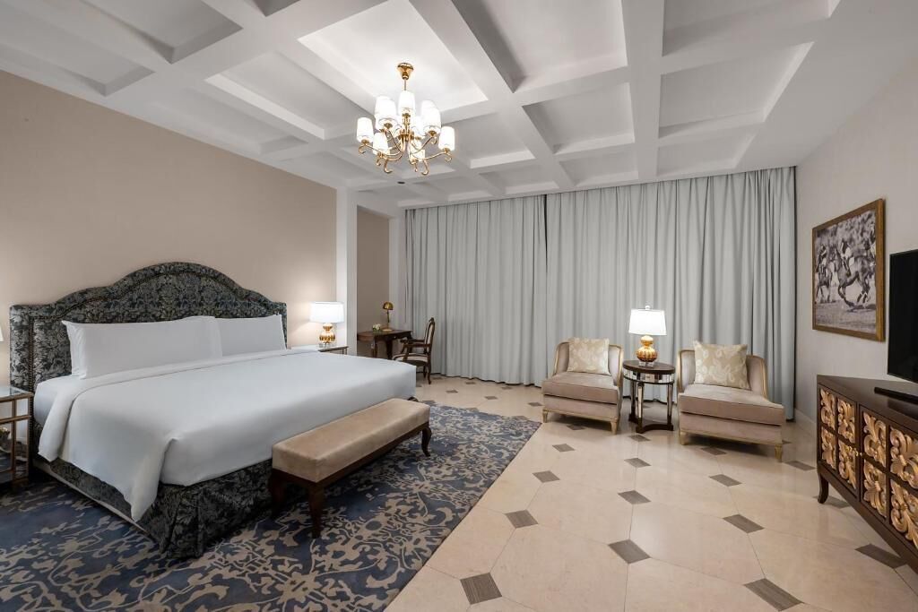 A hotel room with a large bed, two armchairs, a bench, a chandelier, and a television.