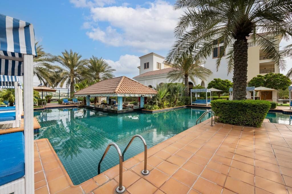 A swimming pool with a tiled deck, lounge chairs, palm trees, and a small gazebo.