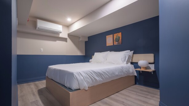 A bedroom with a bed, an air conditioning unit, a bedside table with a lamp, and two wall art pieces. - Hotel Beebop (Toronto)