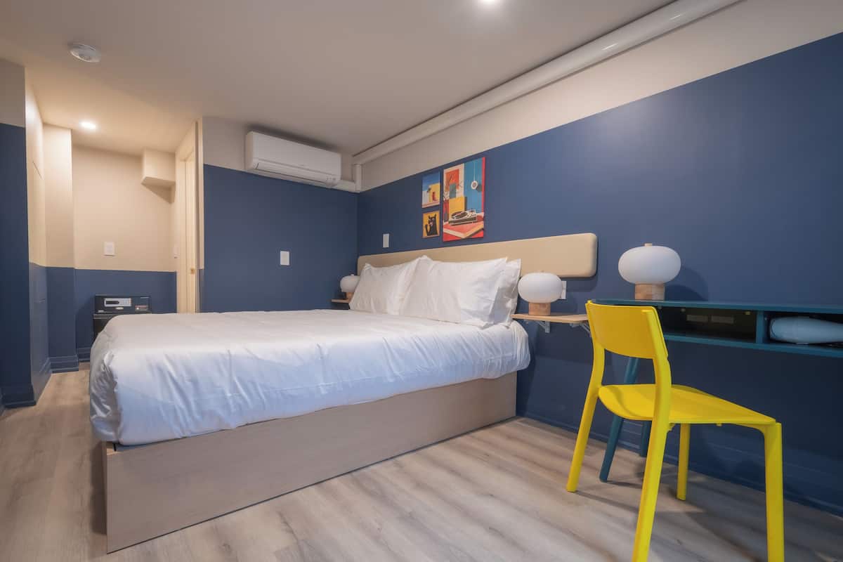 A bedroom with a bed, bedside tables, a desk with a chair, and an air conditioning unit.