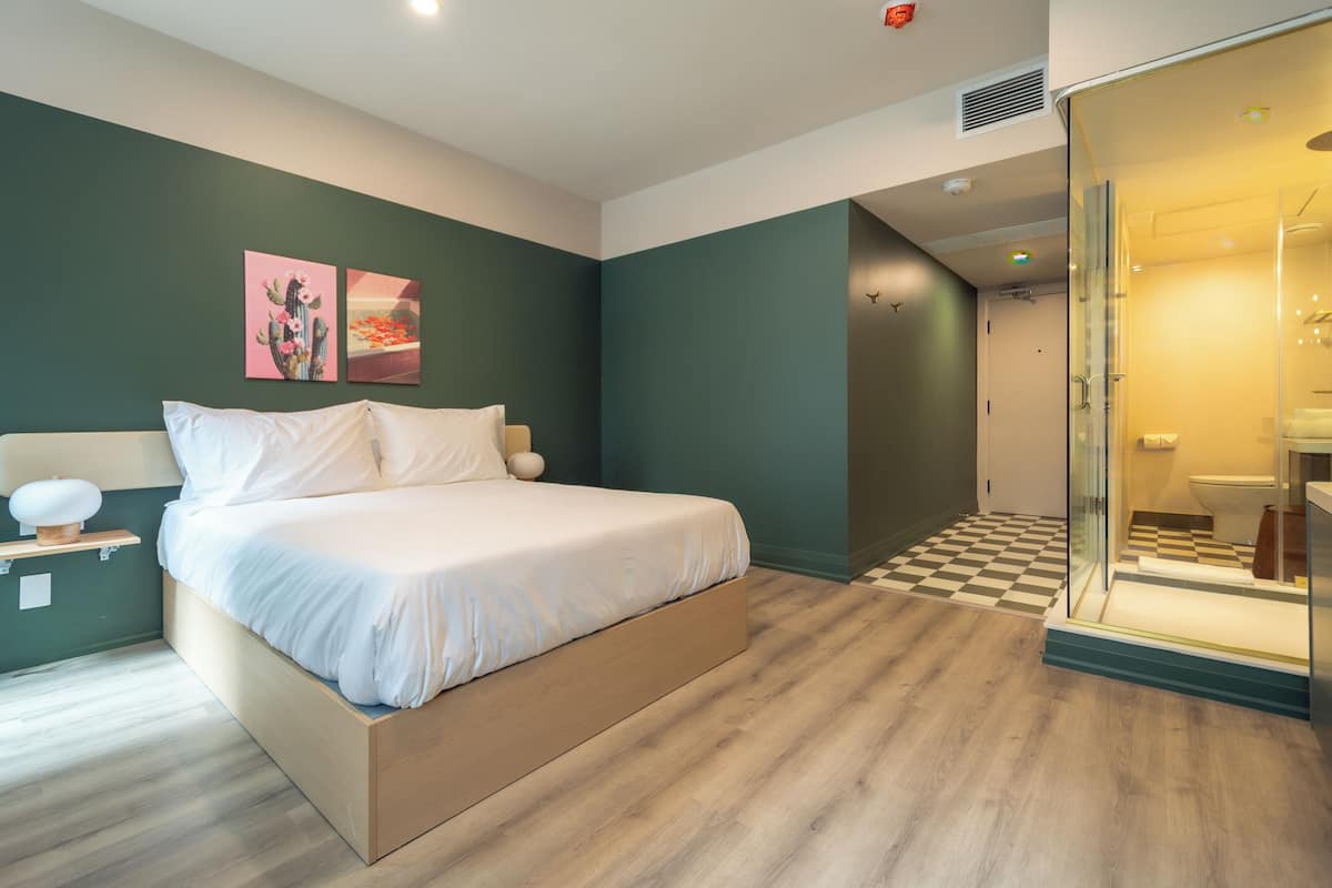 A modern bedroom with a bed, a bedside table, a nightstand, a wall-mounted artwork, and a bathroom visible through a glass door.