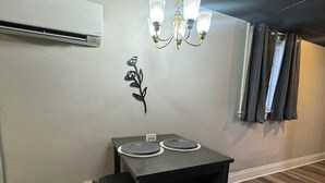 A compact room with a dining table, two chairs, a chandelier, a wall-mounted air conditioner, and a window with curtains. - Cozy 1-bedroom apartment in charming Shepherdsville with AC (Shepherdsville)