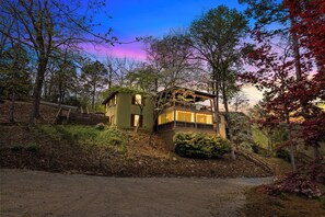 Une maison à deux étages avec un balcon, entourée d’arbres et bordée d’un chemin de terre.
