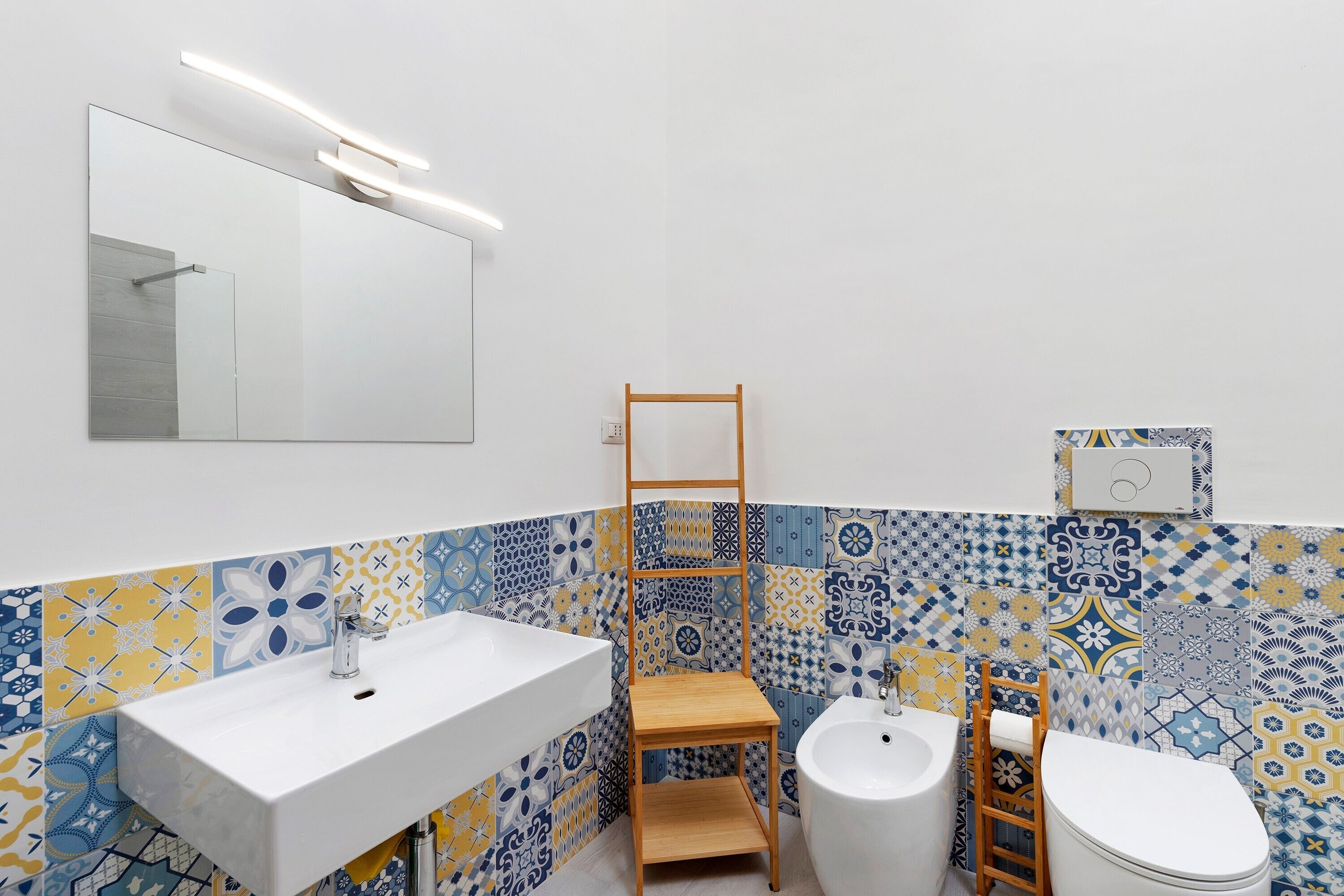 Une salle de bain avec un lavabo blanc, une étagère en bois, des toilettes blanches et des carreaux à motifs.