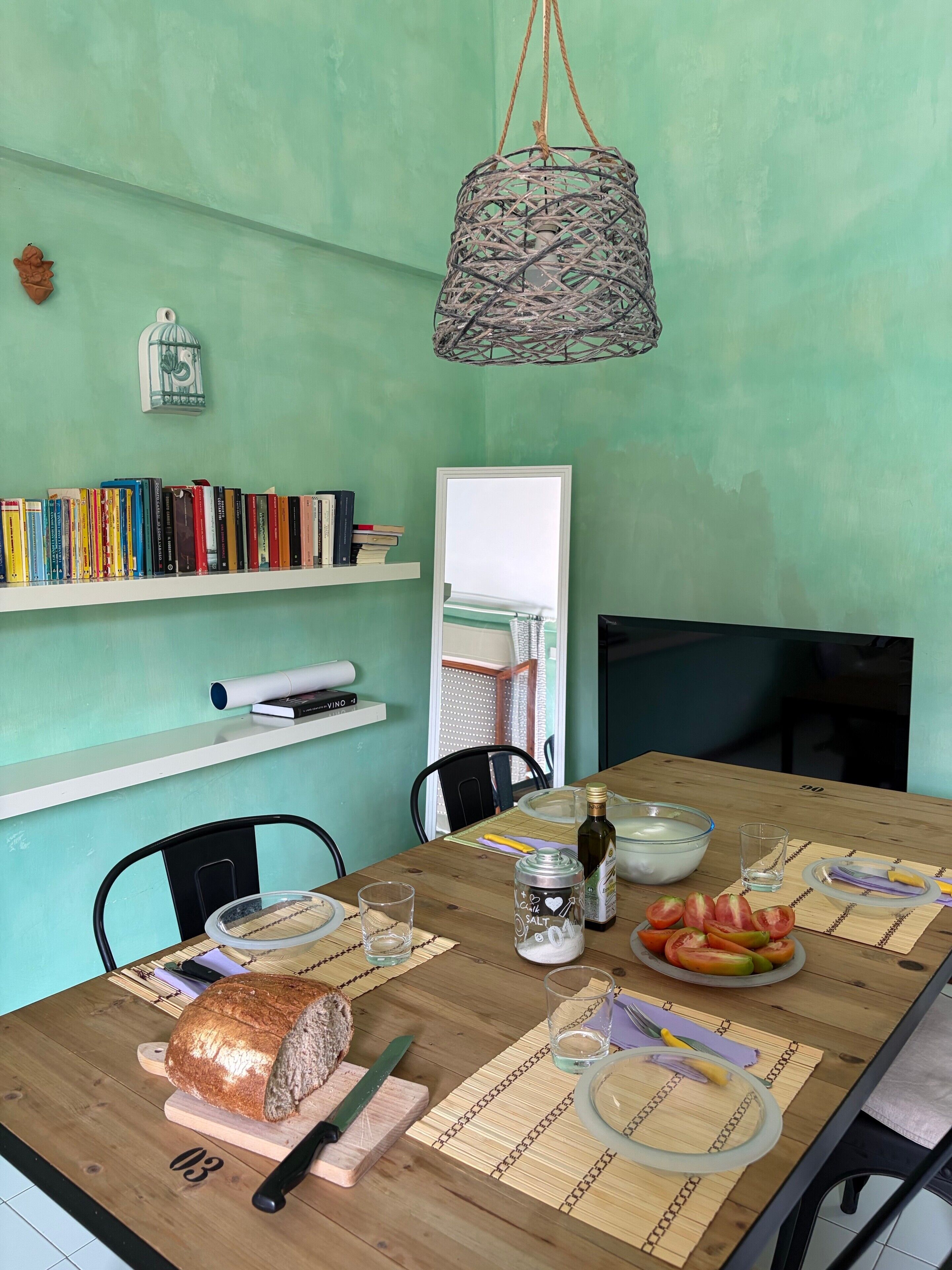 Ein Esstisch mit Brot, einem Messer, einem Salzstreuer, einer Flasche, einer Tomatenscheibe und einem Glas. Es sind Stühle, eine Pendelleuchte und eine Bücherwand mit Büchern zu sehen.