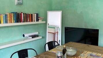 Ein Esstisch mit Brot, einem Messer, einem Salzstreuer, einer Flasche, einer Tomatenscheibe und einem Glas. Es sind Stühle, eine Pendelleuchte und eine Bücherwand mit Büchern zu sehen.