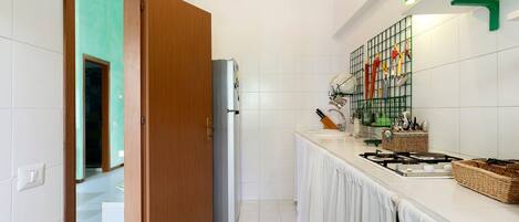 A kitchen with white tiles, a refrigerator, a stove, and a shelf with kitchen utensils.