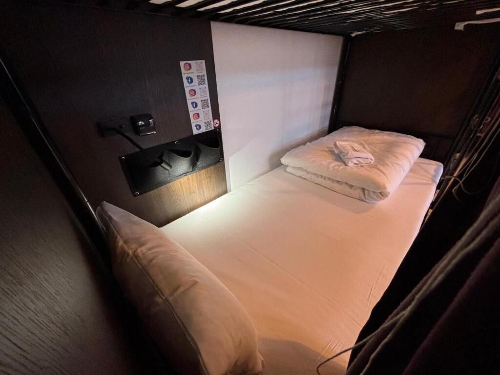 Un lit simple avec un oreiller et une serviette pliée, une petite table de chevet et un panneau de commande fixé au mur, doté de boutons et d’un code QR.