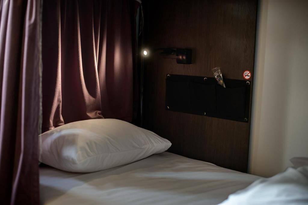 Una cama individual con almohada blanca, cabecera de madera, una lámpara de pared y una puerta con cartel de prohibido fumar.