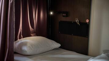 Una cama individual con almohada blanca, cabecera de madera, una lámpara de pared y una puerta con cartel de prohibido fumar.