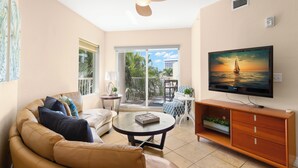 A living room with a beige sofa, a coffee table, a TV on a wooden stand, and a balcony with a view of the ocean. - Crystal Palms 207 (Treasure Island)