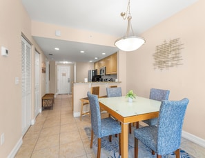 A dining area with a table and chairs, a kitchen in the background, and a hallway leading to a door.